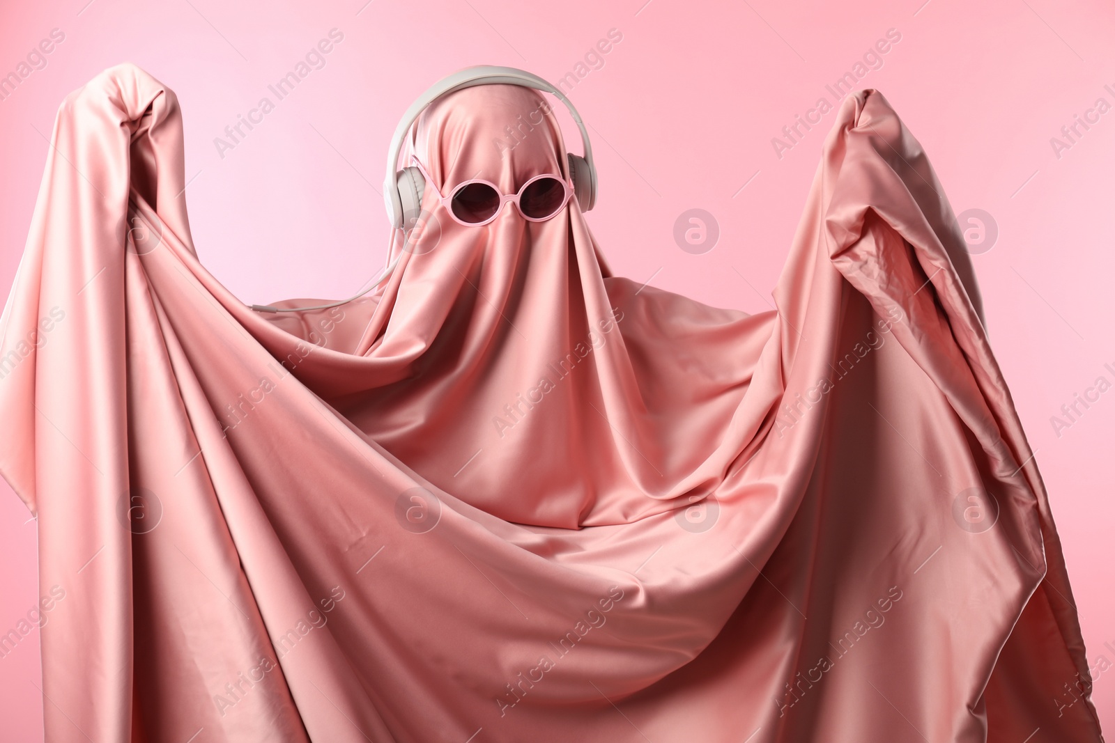 Photo of Glamorous ghost. Woman in sheet with sunglasses and headphones on pink background