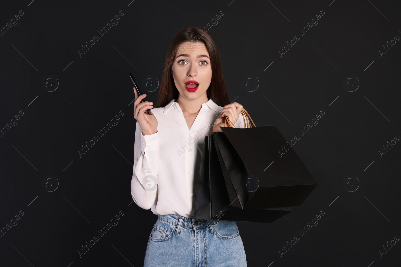 Photo of Surprised young woman with shopping bags and smartphone on black background. Big sale