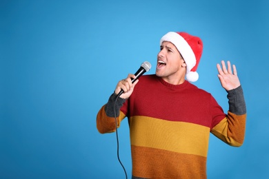 Photo of Emotional man in Santa Claus hat singing with microphone on blue background, space for text. Christmas music