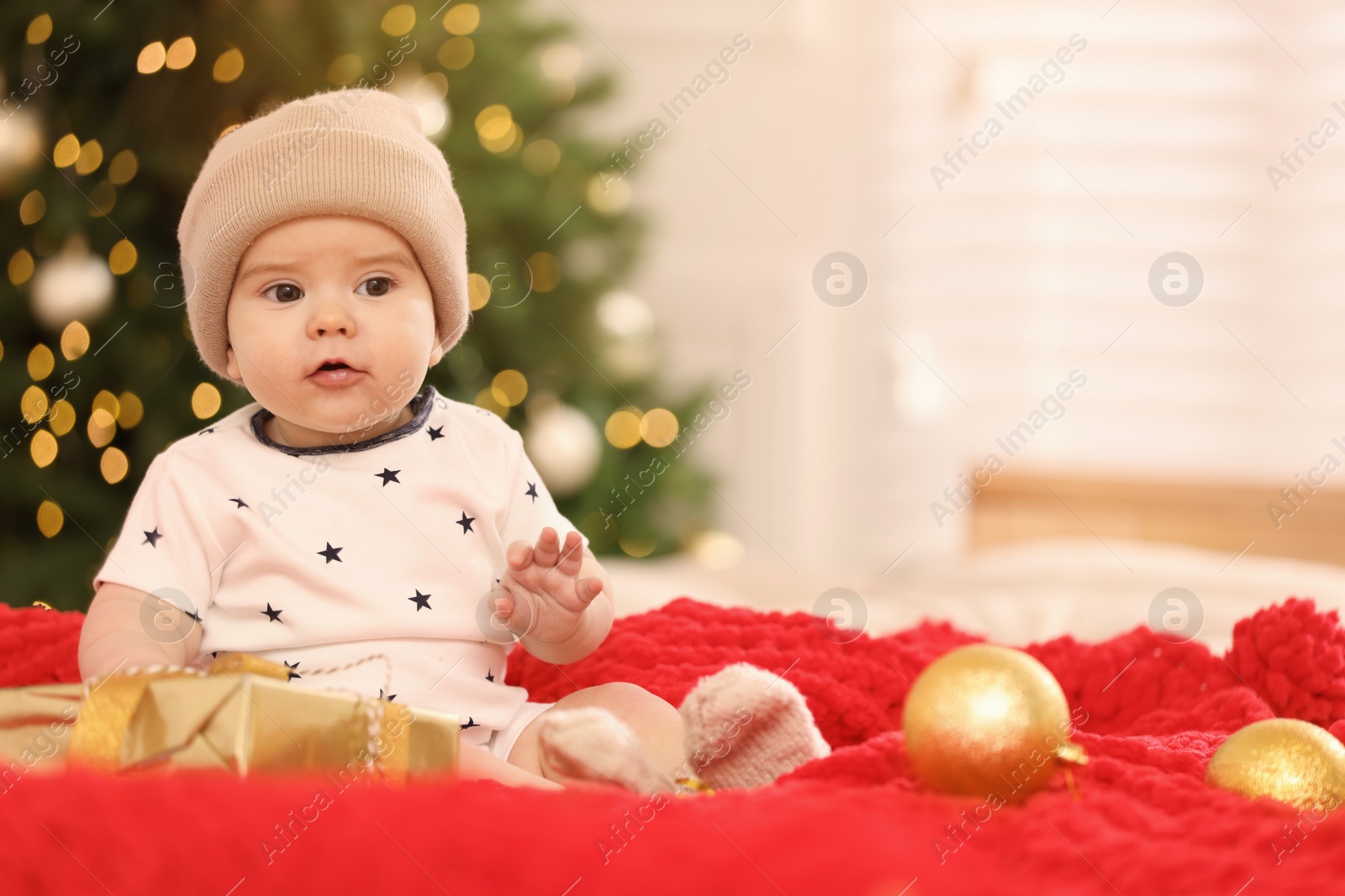 Photo of Cute little baby with Christmas gift on red blanket against blurred festive lights, space for text. Winter holiday