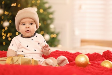 Cute little baby with Christmas gift on red blanket against blurred festive lights, space for text. Winter holiday
