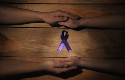 Photo of Man and woman holding hands near purple awareness ribbon on wooden background, top view