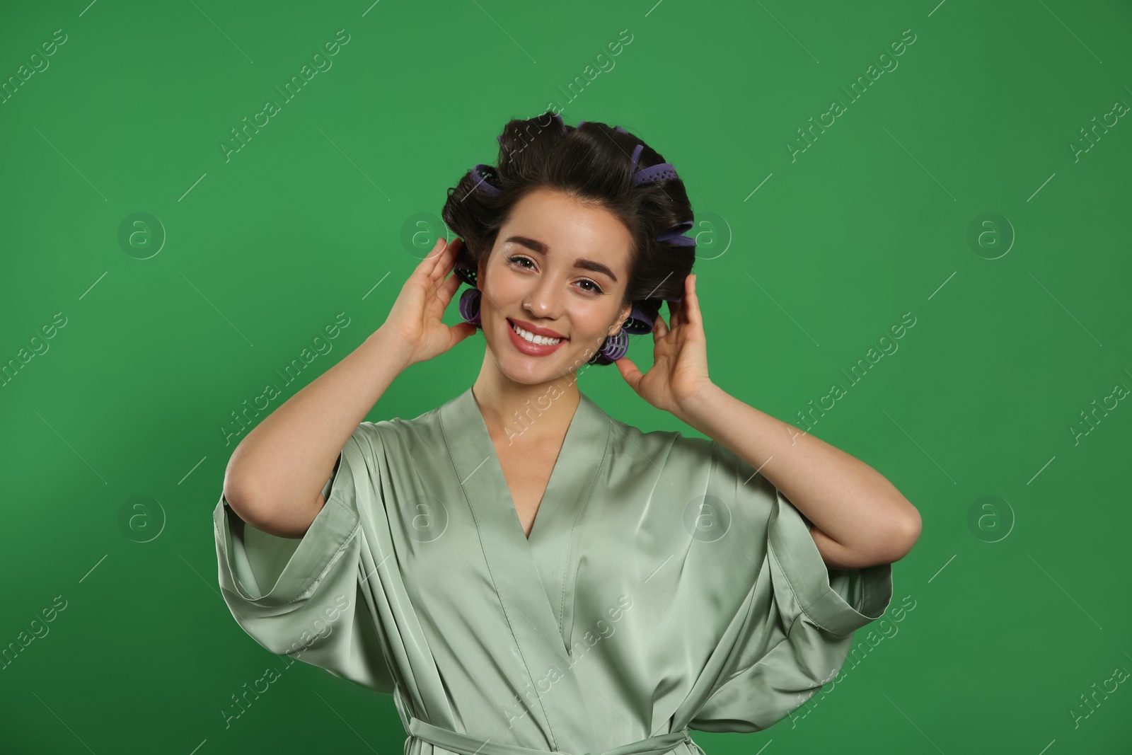 Photo of Happy young woman in silk bathrobe with hair curlers on green background