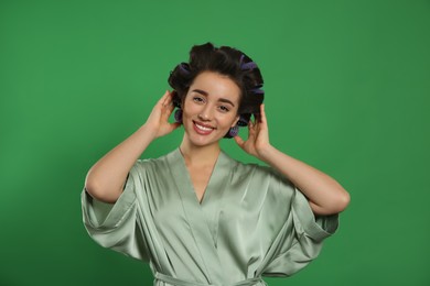 Happy young woman in silk bathrobe with hair curlers on green background