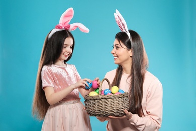 Mother and daughter in bunny ears headbands with basket of Easter eggs on color background