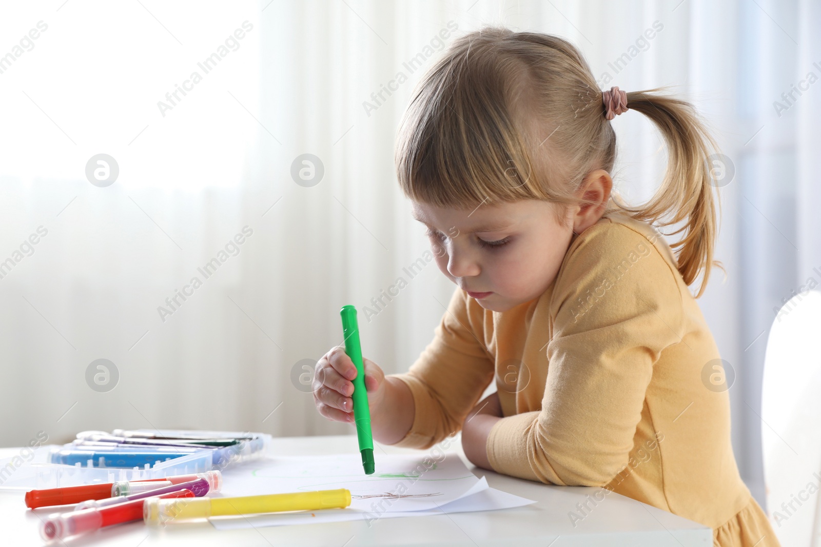 Photo of Cute little girl drawing with marker at white table indoors. Child`s art