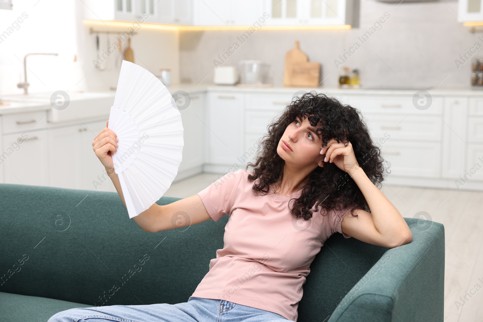 Photo of Young woman waving white hand fan to cool herself on sofa at home