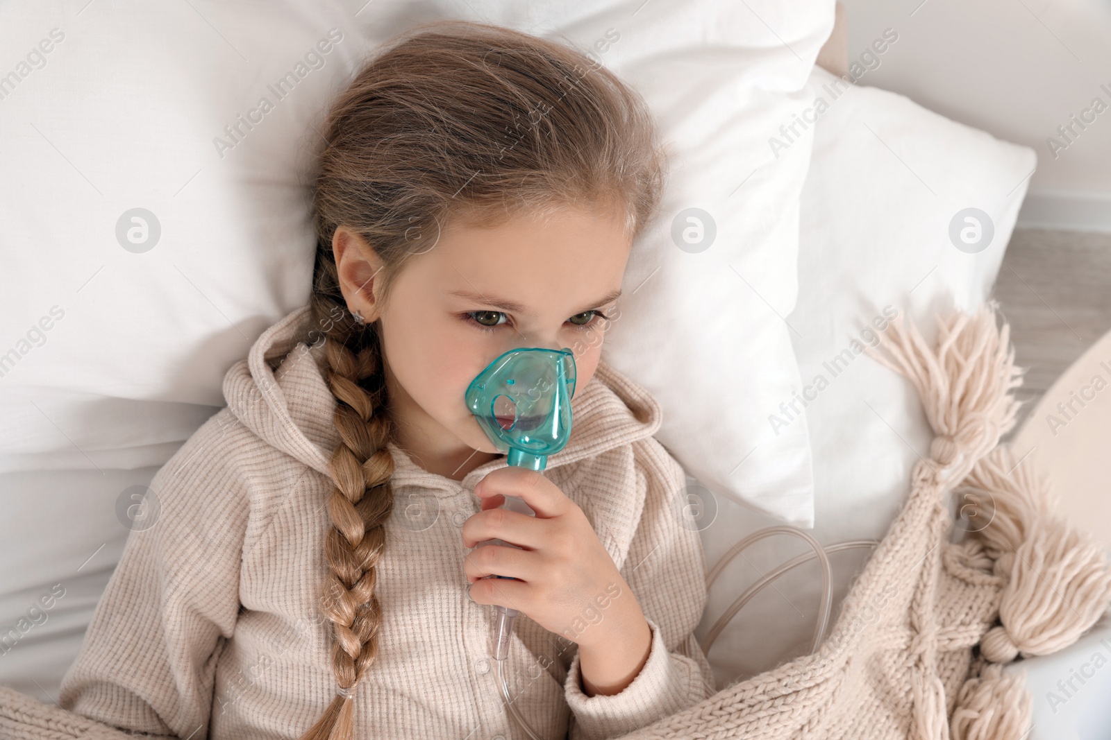 Photo of Little girl using nebulizer for inhalation on bed indoors, top view