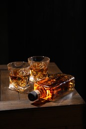 Whiskey with ice cubes in glasses and bottle on wooden crate against black background, closeup