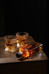 Photo of Whiskey with ice cubes in glasses and bottle on wooden crate against black background, closeup