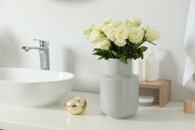 Vase with beautiful white roses and toiletries near sink in bathroom