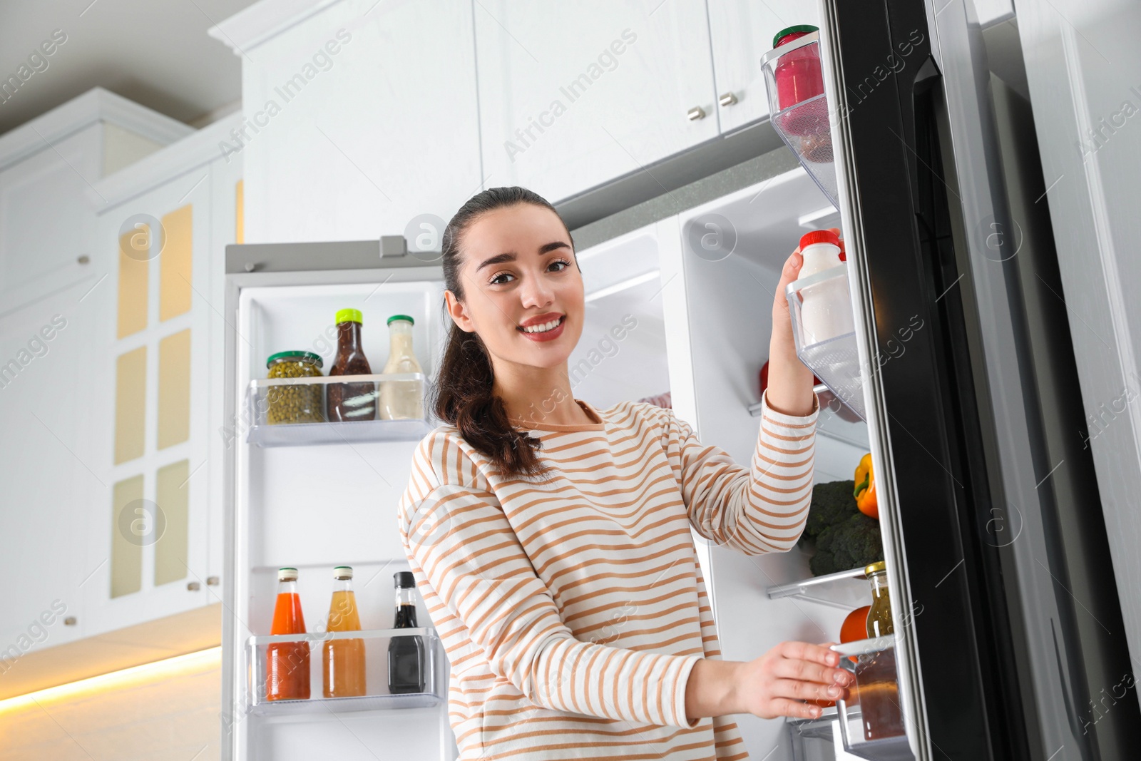 Photo of Young woman taking yoghurt out of refrigerator in kitchen, low angle view