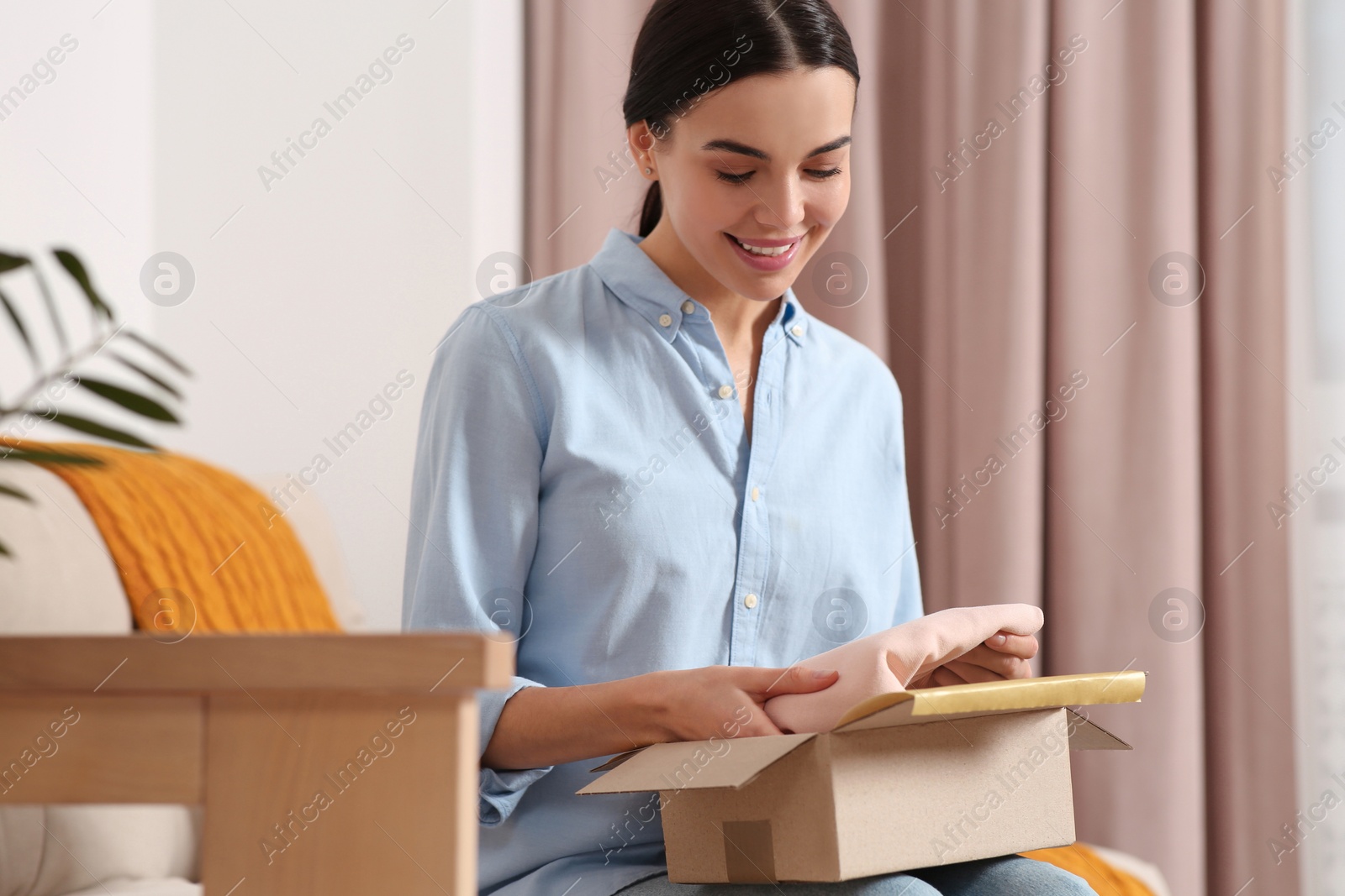 Photo of Happy young woman opening parcel on sofa at home. Internet shopping