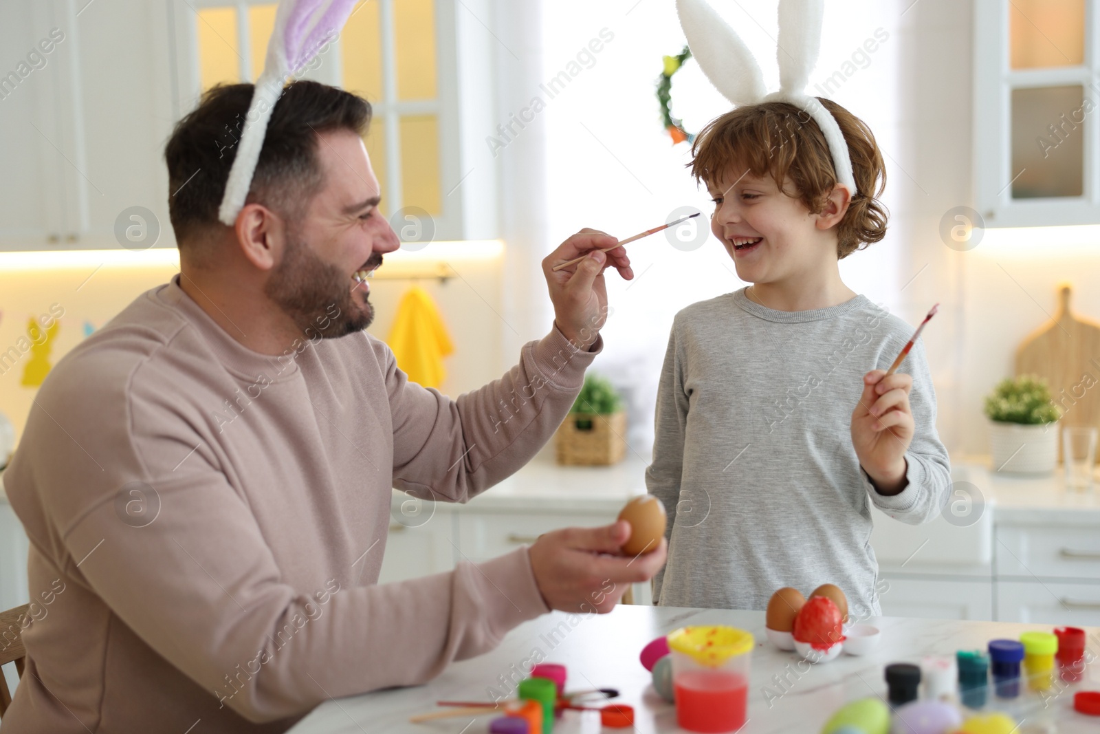 Photo of Easter celebration. Father with his little son having fun while painting eggs at white marble table in kitchen