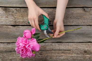 Photo of Woman trimming beautiful pink peonies with secateurs at wooden table, top view