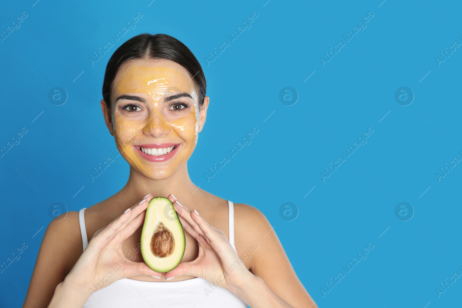 Photo of Young woman with cleansing mask on her face holding avocado against color background, space for text. Skin care