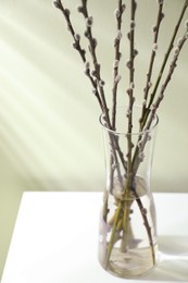 Photo of Glass vase with pussy willow tree branches on white table near light green wall