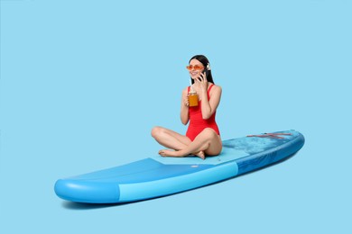 Photo of Happy woman with refreshing drink resting on SUP board and talking on smartphone against light blue background