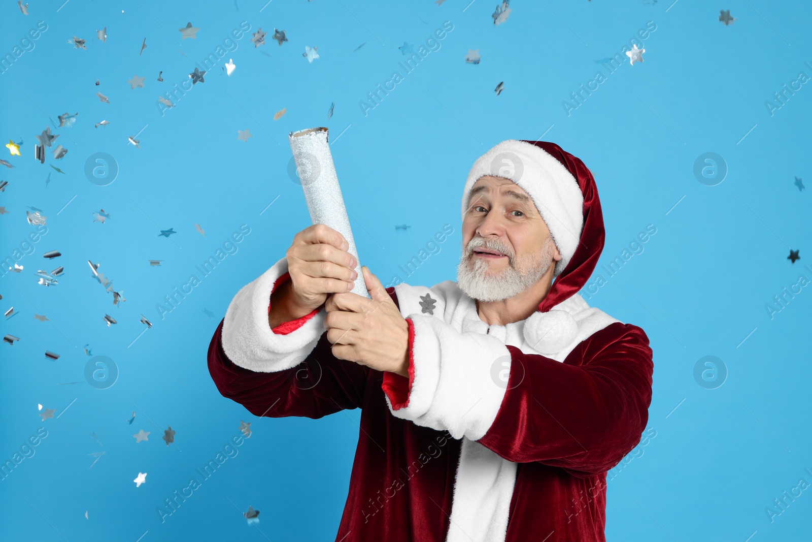 Photo of Man in Santa Claus costume blowing up party popper on light blue background