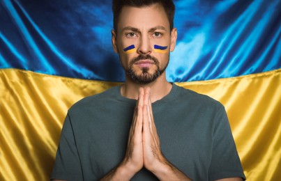 Sad man with clasped hands praying near Ukrainian flag