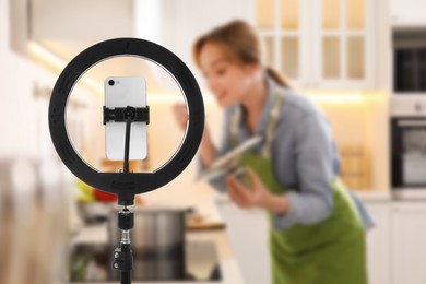 Blurred view of woman tasting food in kitchen, focus on ring light with smartphone