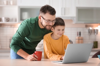 Photo of Little boy and his dad using laptop in kitchen