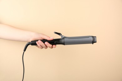 Hair styling appliance. Woman holding curling iron on beige background, closeup