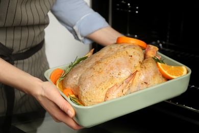 Photo of Woman putting pan with raw chicken and oranges into oven, closeup