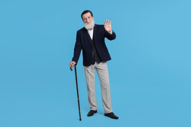 Senior man with walking cane waving on light blue background