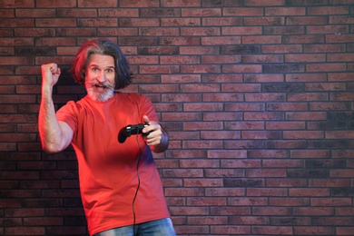 Photo of Emotional mature man playing video games with controller near brick wall. Space for text