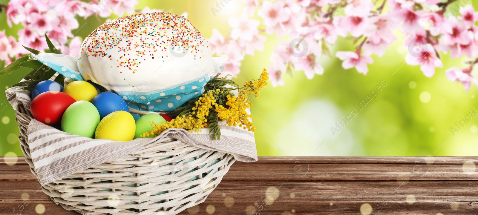 Image of Basket with traditional Easter cake, eggs and flowers on wooden table outdoors, space for text. Banner design