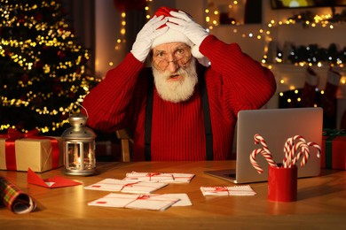 Photo of Emotional Santa Claus at his workplace. Letters and laptop on table in room decorated for Christmas