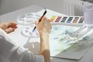 Photo of Woman painting flowers with watercolor at white table indoors, closeup. Creative artwork