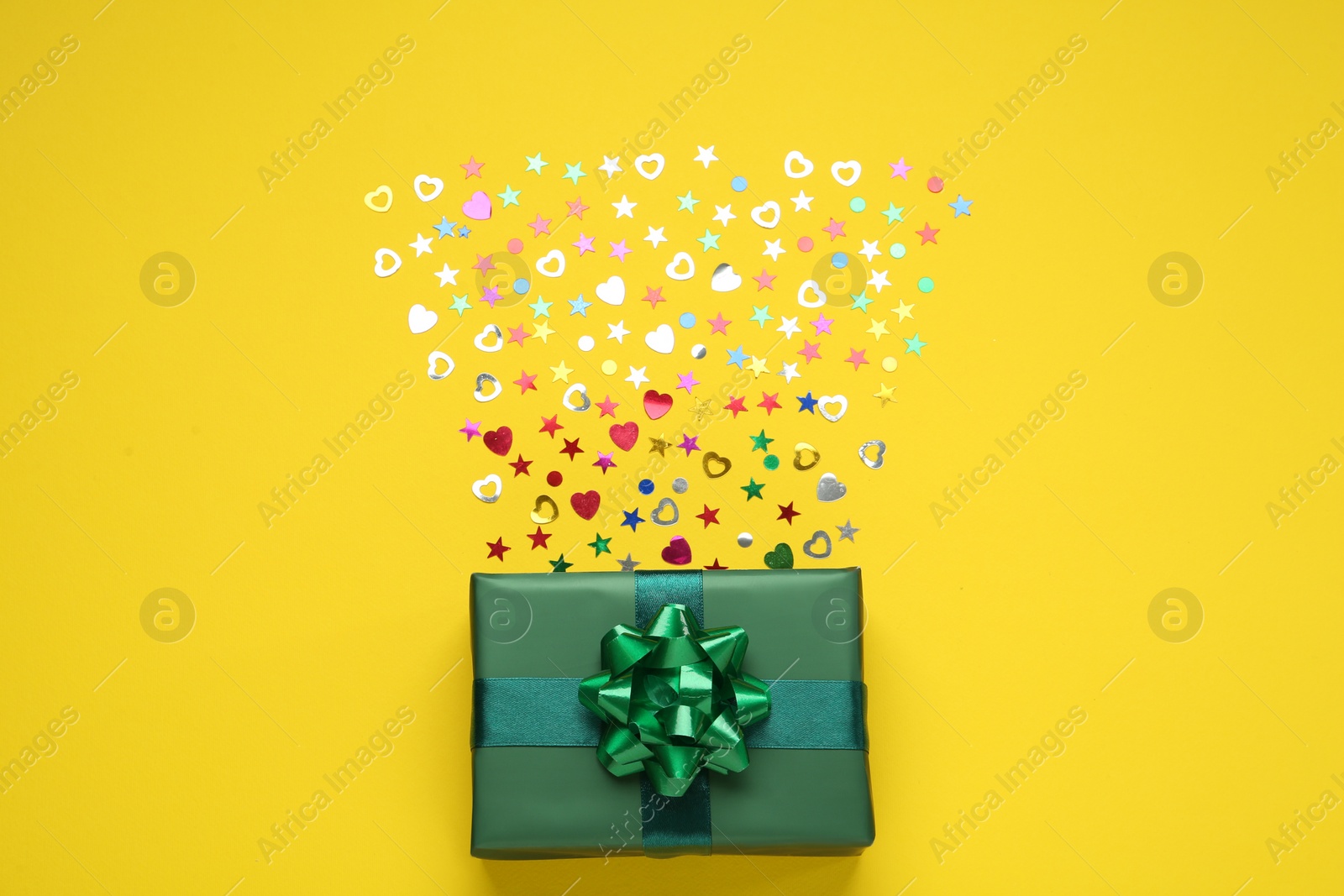 Photo of Green gift box and shiny confetti on yellow background, top view