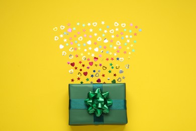 Photo of Green gift box and shiny confetti on yellow background, top view