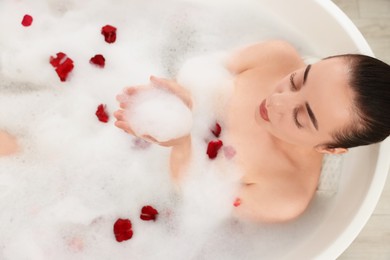 Photo of Woman taking bath in tub with foam and rose petals, top view