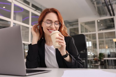 Happy woman with paper cup of coffee working on laptop at white desk in office