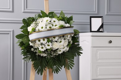 Funeral wreath of flowers on wooden stand and photo frame with black ribbon on white commode indoors