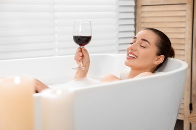 Photo of Happy woman with glass of wine taking bath in tub with foam and rose petals indoors