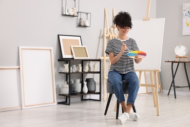 Photo of Young woman with brush and artist`s palette near easel in studio