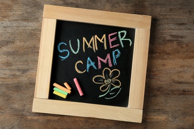 Photo of Small blackboard with text SUMMER CAMP, drawing and chalk sticks on wooden background, top view