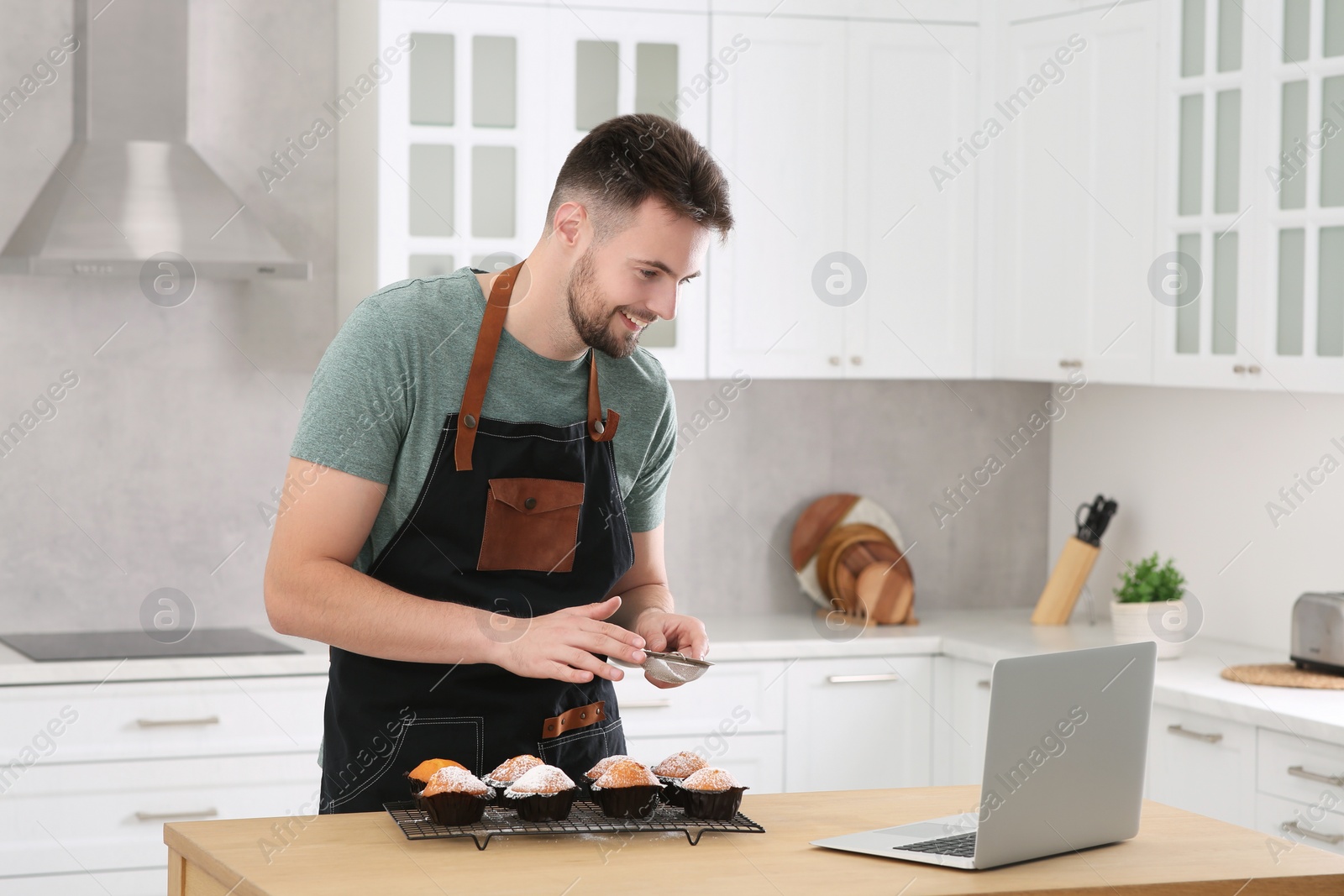 Photo of Man decorating muffins with powdered sugar while watching online cooking course via laptop in kitchen. Time for hobby