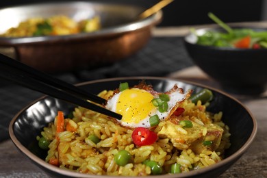 Photo of Taking egg with chopsticks from bowl with tasty rice, meat and vegetables on wooden table, closeup