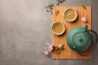 Photo of Traditional ceremony. Cups of brewed tea, teapot and sakura flowers on grey table, flat lay with space for text