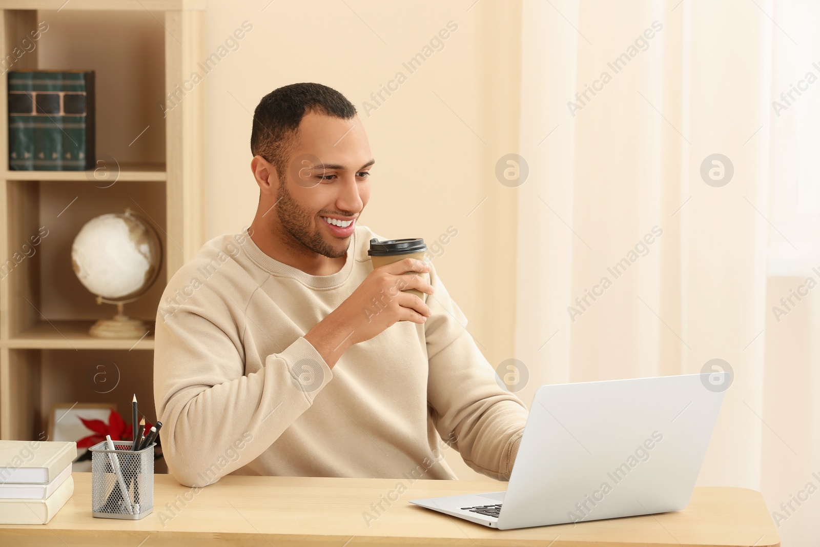 Photo of Smiling African American man with coffee spending time near laptop at wooden table in room