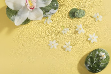 Photo of Beautiful flowers and spa stones in water on pale yellow background, top view