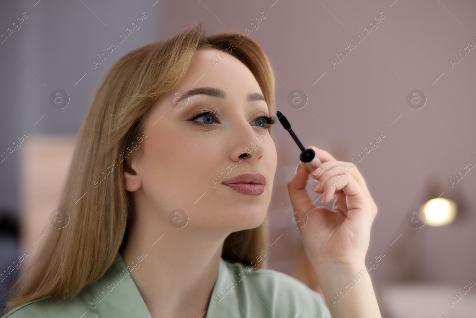 Photo of Reflection of beautiful young woman applying mascara in mirror at home