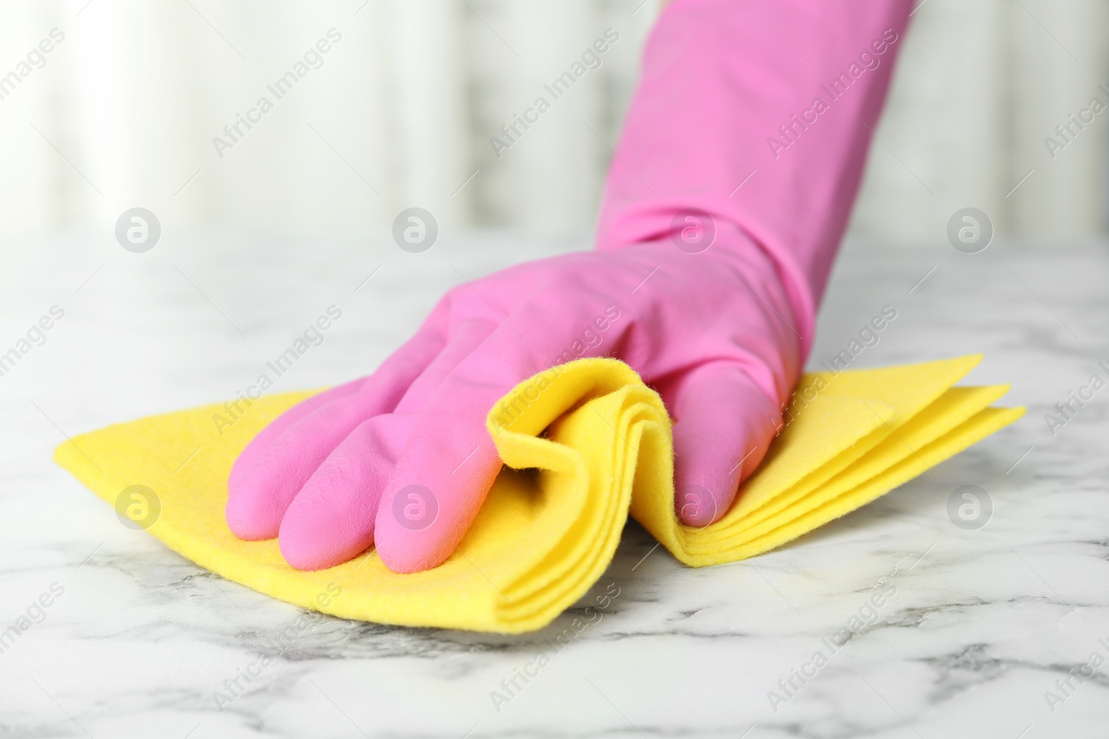 Photo of Woman in gloves wiping white marble table with rag indoors, closeup