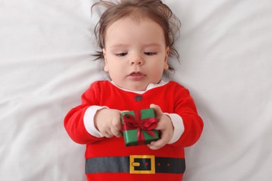Photo of Cute baby wearing festive Christmas costume with gift box on white bedsheet, top view