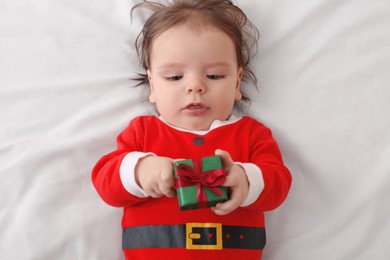 Cute baby wearing festive Christmas costume with gift box on white bedsheet, top view